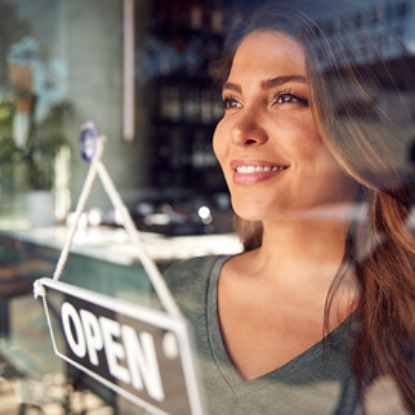 Woman business owner looking through her store window with an open sign in the foreground