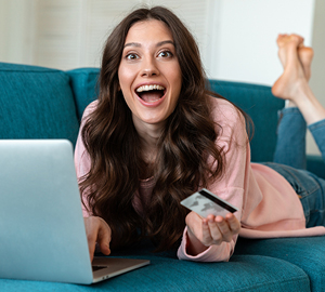 Young female laying on her couch smiling while holding her debit card and working on her laptop.