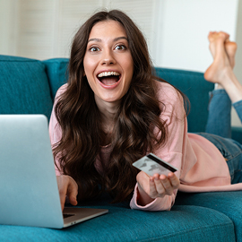 Young woman laying on the couch with her feet crossed holding a debit card working on a laptop