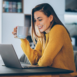 Woman in a yellow sweater holding a cup of coffee while looking at her laptop