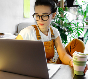 Woman wearing glasses holding a cup of coffee looking at her laptop.